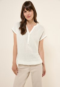 Hvid, kortærmet top med en struktureret stof, V-hals og fem-knap lukning. Kombineret med beige bukser for et afslappet look.