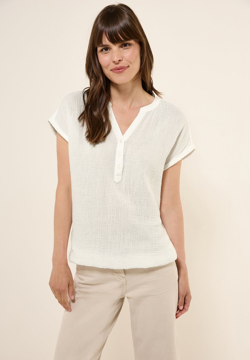 Hvid, kortærmet top med en struktureret stof, V-hals og fem-knap lukning. Kombineret med beige bukser for et afslappet look.