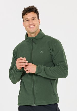 Mand smilende, iført grøn fleecejakke med lynlås og sidelommer, justerer ærmet mod hvid baggrund.