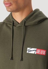 Alkanto žalios spalvos džemperis su raišteliu, turintis baltą ir raudoną logotipo pleistrą su užrašu "Tommy JEANS", pagamintas iš minkštos medžiagos.