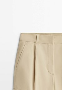 Shorts beiges sur mesure avec passants pour ceinture, poches latérales et une couture avant, au design épuré et minimaliste sur fond blanc.