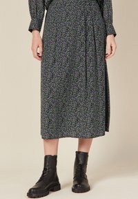 Schwarzer Midirock mit Blumenmuster und lila sowie grünen Akzenten, in einem fließenden Design mit plissiertem Detail. Kombiniert mit schwarzen Schnürstiefeln.