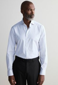 Chemise blanche avec des rayures verticales bleu clair, à manches longues, col pointu et fermeture à boutons. Associée à un pantalon noir.