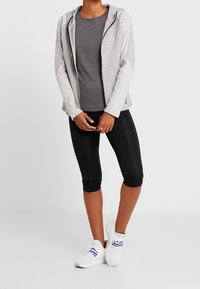 Světlé šedé mikina na zip s kapucí, doplněná tmavě šedým tričkem s krátkým rukávem, černými tříčtvrtečními legínami a bílými sportovními botami.