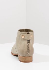 Beiger Knöchelstiefel mit perforiertem Design, vertikalem Reißverschluss und rundem goldfarbenem Akzent, flachem Holzabsatz und weicher Lederstruktur.