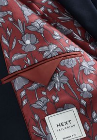 Burgunderfarbener Blumen-Einstecktuch mit grauen Blumen und grünen Blättern, einem kontrastierenden roten Saum und einem Etikett mit der Aufschrift "NEXT TAILORING SKINNY FIT."