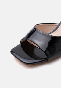 Sandale à bout ouvert en cuir verni noir avec avant carré et large sangle sur les orteils, sur fond beige.