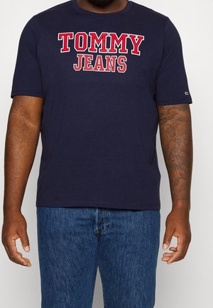 Uomo che indossa una t-shirt blu navy "Tommy Jeans" con scritte rosse e bianche e jeans blu, mostrando un tatuaggio sull'avambraccio destro.