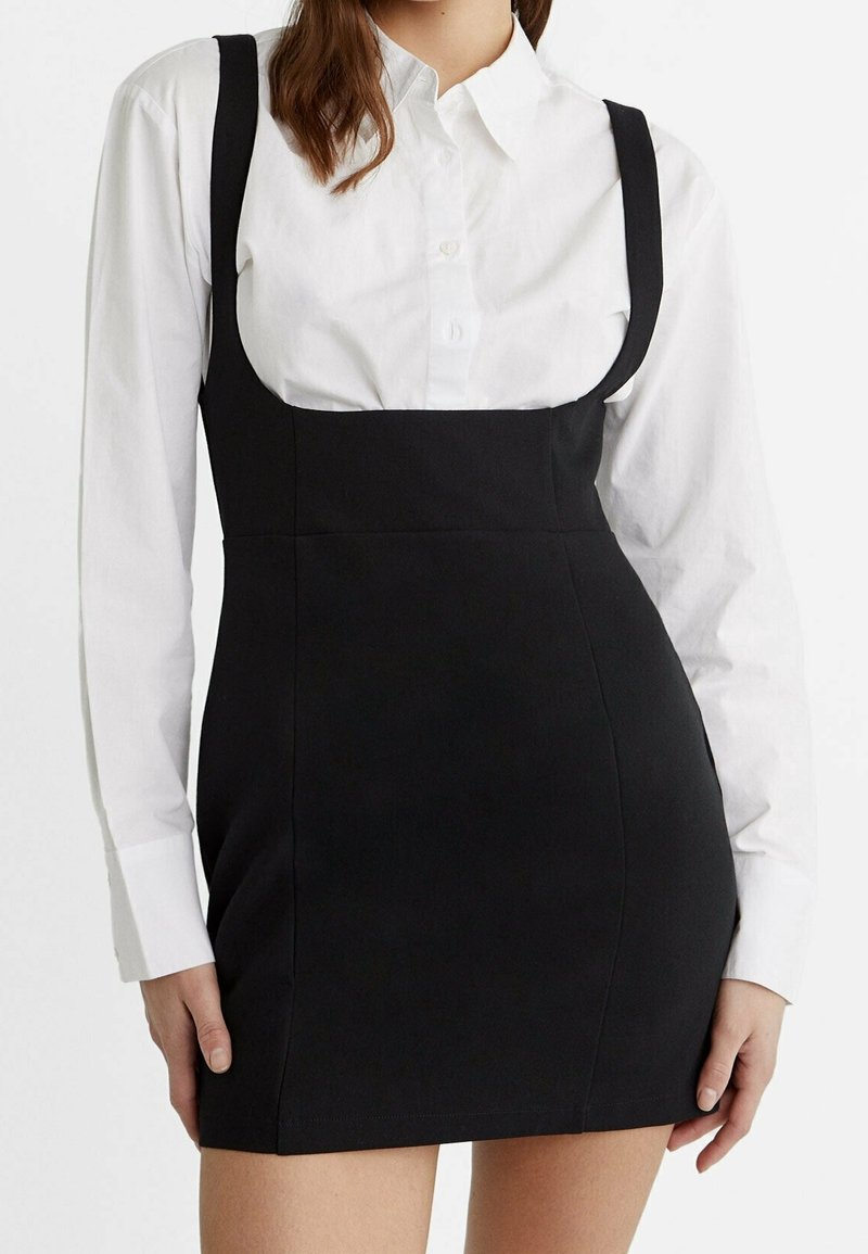 Femme portant une chemise blanche boutonnée sous une robe mini noire ajustée sans manches, montrée des épaules jusqu'au milieu des cuisses.