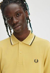 Polo giallo con finitura testurizzata, dotato di un bottone a due fori, strisce a contrasto sul colletto e un piccolo logo nero sul petto.