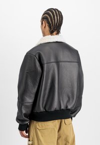 Svart skinnjakke med saueskinnskrage, teksturert overflate, ribbestrikkede ermer og linning. Stilt i en oversized design, bakview vist.