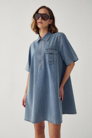 Žena oblečená v oversized rifľových šatách s krátkym rukávom, predným zipsom a náprsnom vreckom, s veľkými čiernymi slnečnými okuliarmi, stojaca na jednoduchom pozadí.