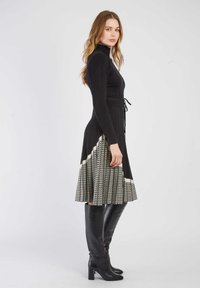 Schwarzes Langarmkleid mit geripptem Oberteil, tailliertem Bund und plisseeartigem Rock im Hundezahn-Muster. Kombiniert mit knielangen schwarzen Stiefeln.