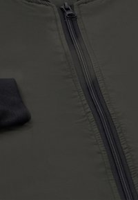 Sort jakke lavet af et glat, fyldigt materiale med en frontlynlås og en strikket manchet. Stoffet er mørkt og mat med subtile teksturdetaljer.