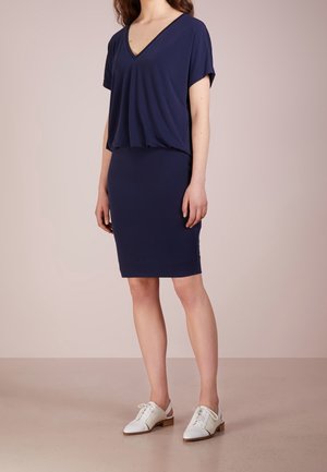 Vrouw draagt een marineblauwe korte mouwen V-hals jurk met een aansluitende rok en witte veterschoenen met lage hak tegen een effen achtergrond.