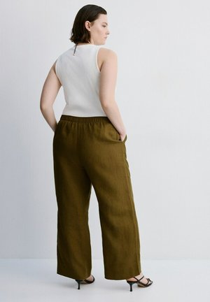 Mujer de pie con las manos en los bolsillos, vestida con una blusa blanca sin mangas, pantalones anchos de color verde oliva y sandalias negras de tacón sobre un fondo blanco.