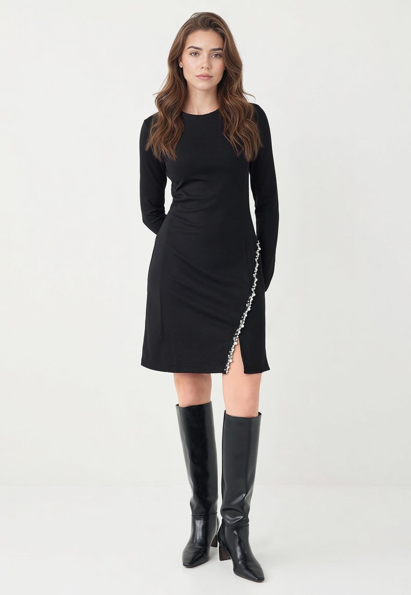 Czarna sukienka z długim rękawem z rozcięciem po boku i dekoracyjnymi białymi guzikami wzdłuż lewego szwu, w zestawie z czarnymi, wysokimi do kolan skórzanymi kozakami.