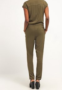 Femme portant une combinaison courte à manches courtes de couleur vert olive avec des poches, se tenant avec les mains dans les poches, chaussée de talons hauts noirs.