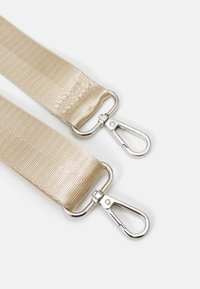 Beige nylonrem med en texturerad yta, utrustad med två metallsvängkrokar. Krokarna har en glänsande silverfärg och en fjädrande mekanism.