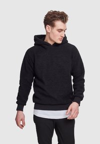 Felpa nera in pile con superficie testurizzata, design con cappuccio, polsini e vita a coste, indossata sopra una camicia bianca a strati e pantaloni neri.
