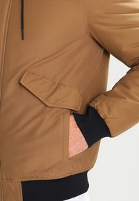 Brown padded jacket with a large flap pocket, ribbed black cuffs, smooth texture, and drawstring hood.