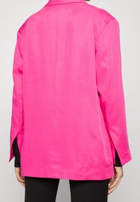 Růžový blejzr s volným střihem, vyrobený z hladkého materiálu. Má široké rukávy a boční rozparky pro snadný pohyb.