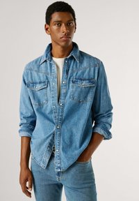 Světle modrá džínová košile s dvěma náprsenkami, klasickým límcem a zapínáním na knoflíky. Dlouhé rukávy jsou ohrnuté na manžetách, nošená přes bílou košili.