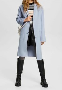 Valkoinen villakangastakki suoran leikkauksen kanssa, leikatut läpät ja kolme nappia; yhdistetty raidalliseen neuleeseen, nahkashortseihin ja polvisukkiin mustissa saappaissa.