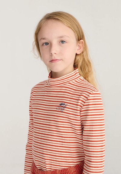 Jeune fille aux longs cheveux blonds portant un col roulé rayé rouge et blanc avec un petit motif floral sur la poitrine.