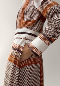 Robe à motifs avec des designs géométriques en orange, marron et blanc. Comprend une taille et une poche latérale, avec un tissu texturé.