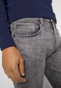 Šedé džínové kalhoty s vybledlým povrchem, které mají přední kapsu s nenápadným logem a kontrastním šitím. Viditelná tmavě modrá košile s dlouhým rukávem.