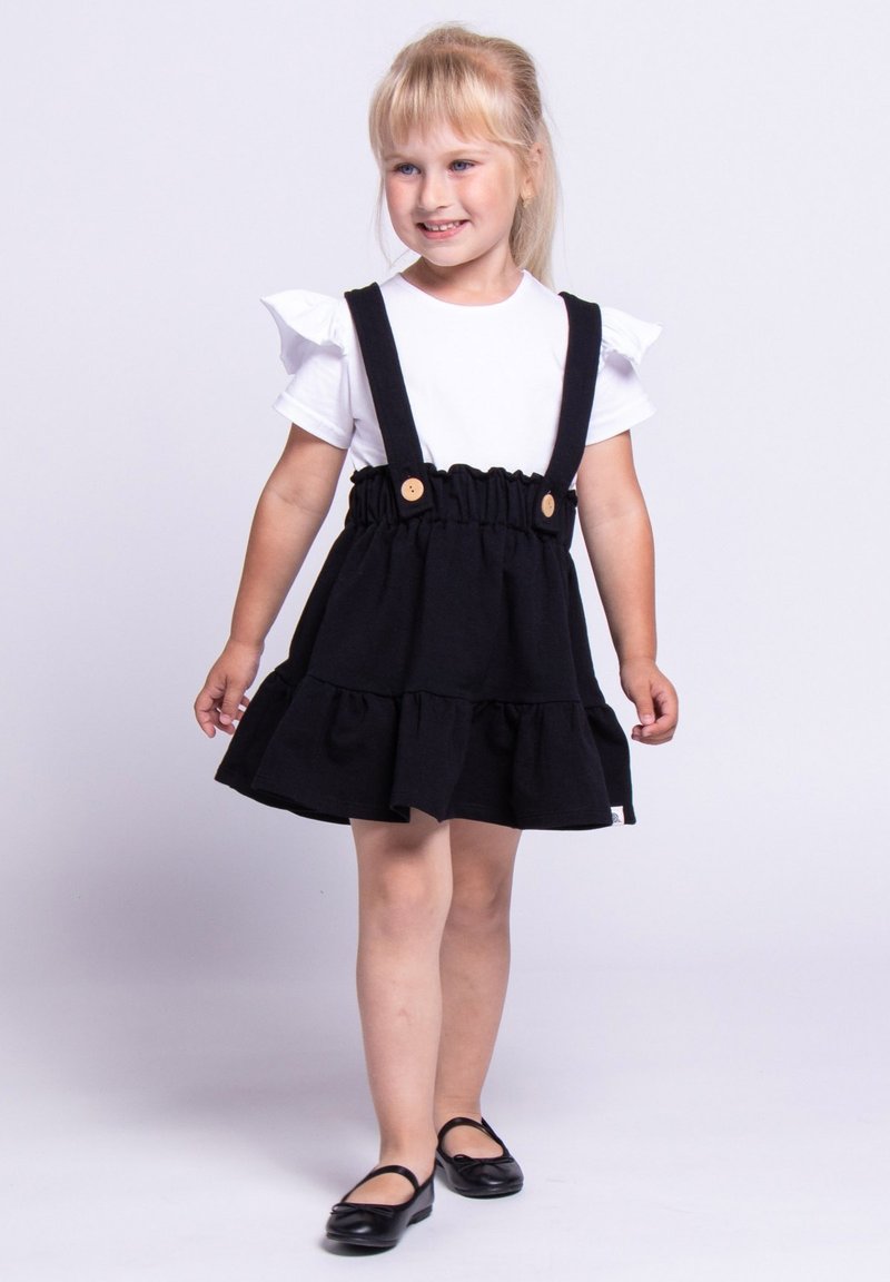 Czarna bawełniana sukienka z falbaniastym dołem, regulowanymi ramiączkami i drewnianymi guzikami. Zestawiona z białą koszulą z krótkim rękawem i czarnymi butami.