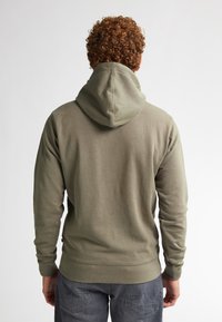 Felpa verde oliva con una finitura testurizzata, dotata di cappuccio con cordoncino e polsini a coste. Sul retro presenta una vestibilità morbida.