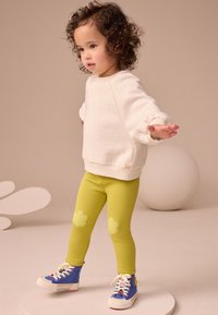 Weiß gesprenkeltes Sweatshirt, grüne Leggings mit Wolkenapplikationen, blaue Turnschuhe mit Regenbogenakzenten, Kind posiert vor neutralem Hintergrund.