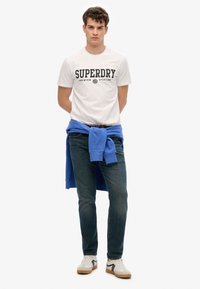 Hvid T-shirt med "SUPERDRY" print, blå sweatshirt bundet om taljen, mørke jeans og beige sneakers med sorte accenter.