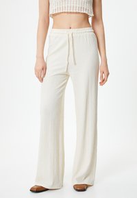 Pantalons à larges jambes, couleur crème, en tissu texturé avec une taille élastique et un cordon de serrage. Associés à des chaussures plates brunes.