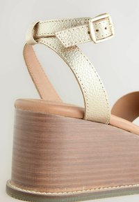 Ék alakú szandál aranyszínű, texturált pánttal, barna bőr talprésszel és fából készült, rétegelt kialakítású sarokkal. Csattal záródó pánt.