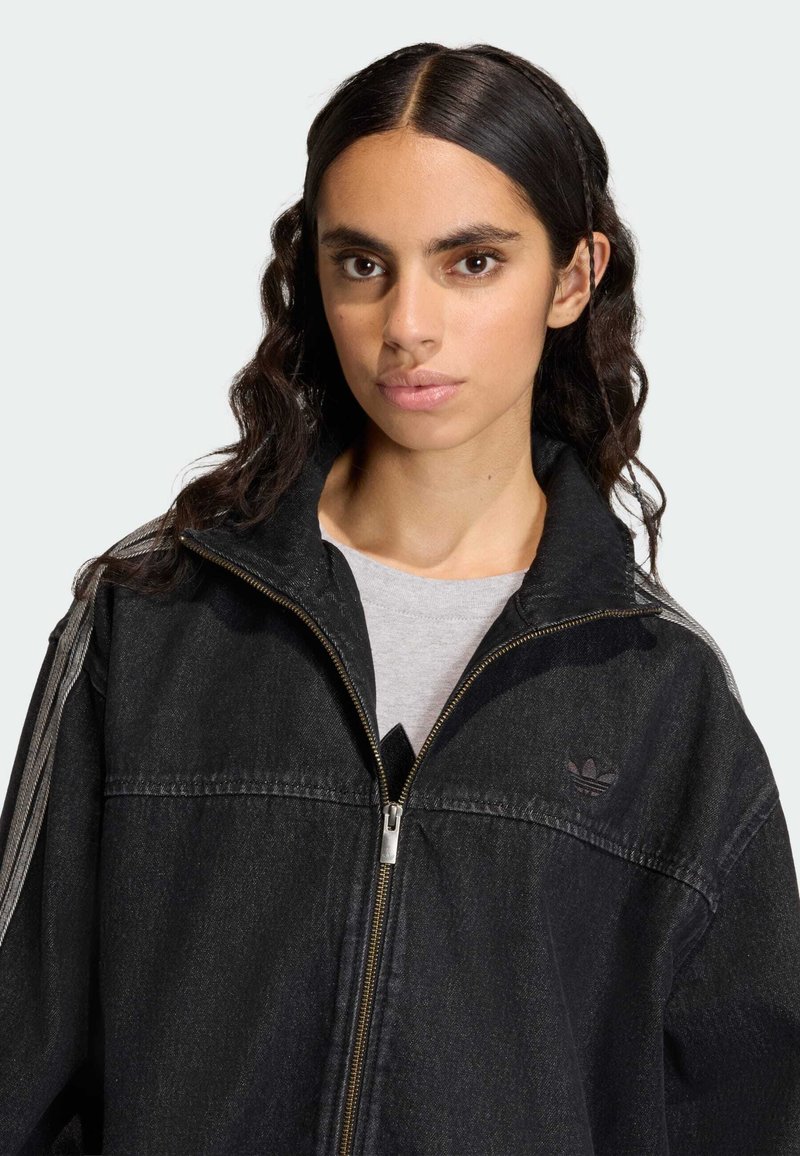 Black denim jacket with a zip closure, high collar, and subtle logo. Features gray stripes on shoulders and a relaxed fit.