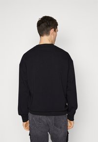 Felpa nera con collo tondo e maniche lunghe, caratterizzata da una texture liscia. Il modello indossa jeans grigi a gamba dritta.