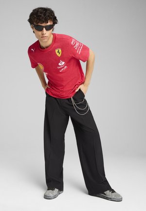 Czerona koszulka Puma z logo Ferrari i nadrukami sponsorów; czarne spodnie z szerokimi nogawkami z akcentami w postaci srebrnego łańcucha; szare sneakersy z czarnymi sznurowadłami.