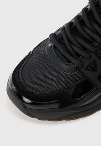 Chaussure de sport noire avec une tige en maille texturée, des accents synthétiques brillants et de larges lacets. Elle présente un bout arrondi et une semelle rembourrée.