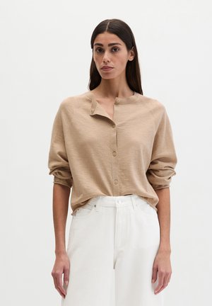 Frau trägt einen beigefarbenen, geknöpften Cardigan mit hochgekrempelten Ärmeln und eine hoch taillierte weiße Hose, steht vor einem einfachen hellen Hintergrund.