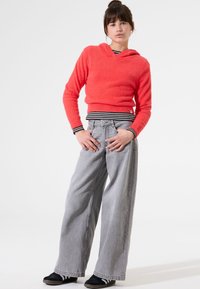 Sudadera roja esponjosa con cuello en V, combinada con pantalones vaqueros anchos de denim grises. Zapatos negros con rayas blancas. Fondo blanco.