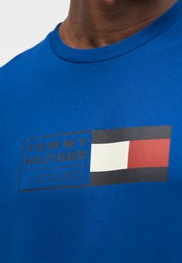 Μπλε βαμβακερό μπλουζάκι με μαύρο ορθογώνιο λογότυπο που φέρει το κείμενο "TOMMY HILFIGER", μία λευκή ράβδωση και ένα κόκκινο-λευκό τετράγωνο διακοσμητικό στοιχείο.