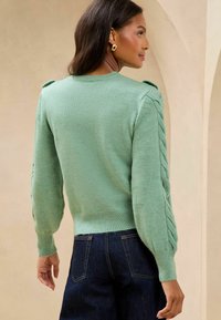 Pulover tricotat de culoare verde deschis, cu mâneci cu model tip cablu, tiv cu nervuri și guler rotund, asociat cu pantaloni din denim de culoare albastru închis.