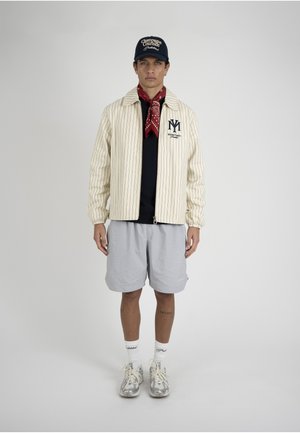 Mladý muž, který má na sobě krémové sako s proužky, červenou bandanu, námořnickou čepici, světle šedé kraťasy, bílé ponožky a stříbrné tenisky na jednobarevném pozadí.