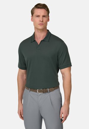 Homme portant un polo manches courtes vert foncé rentré dans un pantalon gris clair avec une ceinture tressée marron, debout devant un fond uni.