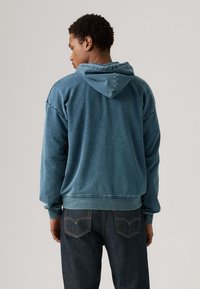 Blå hettegenser med en avslappet passform og ribbestrikkede mansjetter, parret med mørkejeans. Materialet ser mykt og litt teksturert ut.