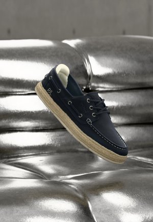 Scarpa da barca blu navy con cuciture bianche e suola in corda beige, che galleggia sopra un divano argentato brillante trapuntato.