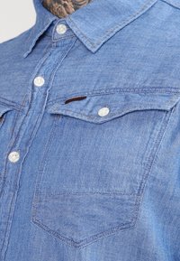 Lichtblauwe denim shirt met een puntkraag, knopen-sluiting, en twee borstzakken met witte knopen en contrasterende stiksels.